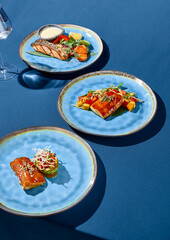 A set of Asian-style dishes featuring salmon, cod, and pike perch, presented on blue ceramic plates against a monochromatic blue background with sharp shadows