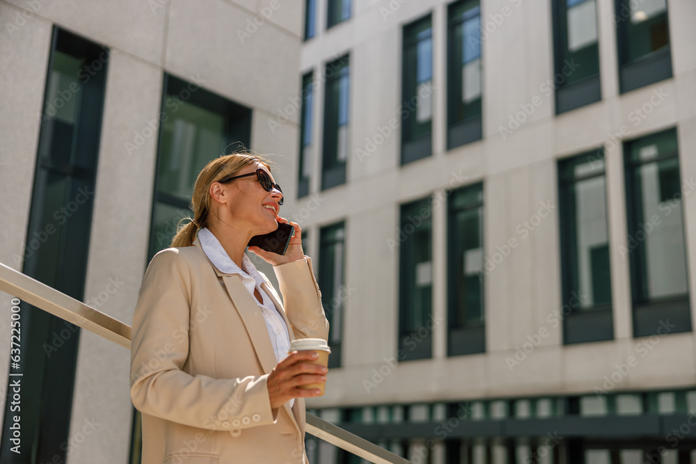 Woman manager talking phone whiile standing on modern building background and drinking coffee