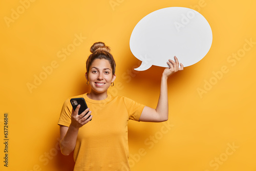 Waist up shot of good looking young European woman with hair bun holds mobile phone and blank communication bubble for your advertising content wears casual t shirt isolated over yellow background.