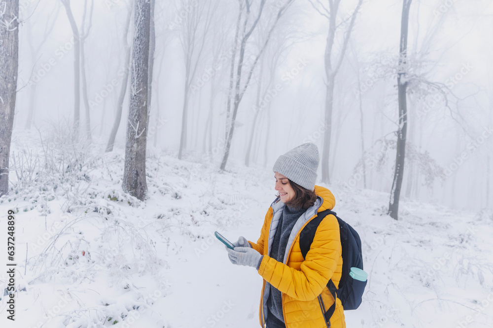 Young caucasian woman in a yellow jacket and hat walking in a snowy forest with a smartphone in her hands