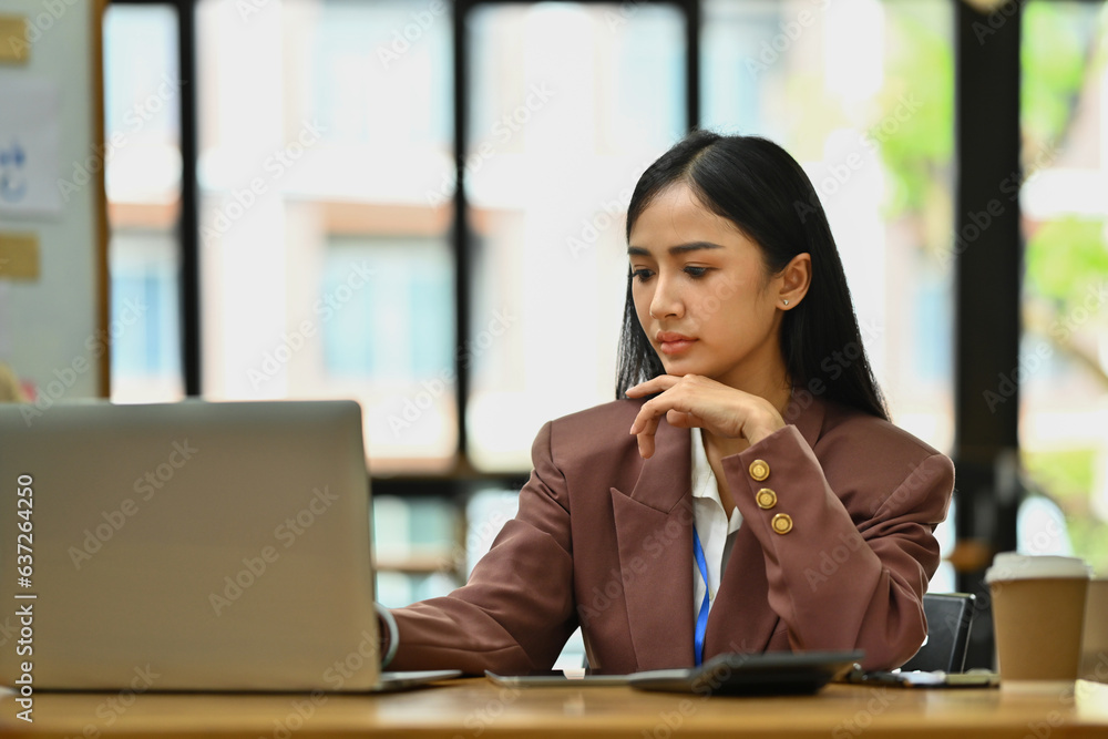 Serious Asian businesswoman sitting at desk with laptop, working with financial accounting documents. Serious Asian businesswoman sitting at desk with laptop, working with financial accounting documents.