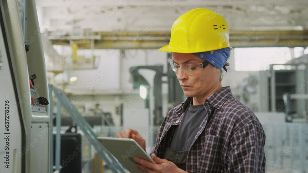 Tilt up shot of female worker in hardhat and safety glasses operating industrial machine with digital tablet in stone cutting factory Tilt up shot of female worker in hardhat and safety glasses operating industrial machine with digital tablet in stone cutting factory
