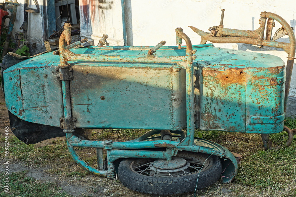 old, rusty, retro green color heavy iron classic motorcycle sidecar for passenger lies on its side with one old wheel on the ground near a white wall on the street during the day