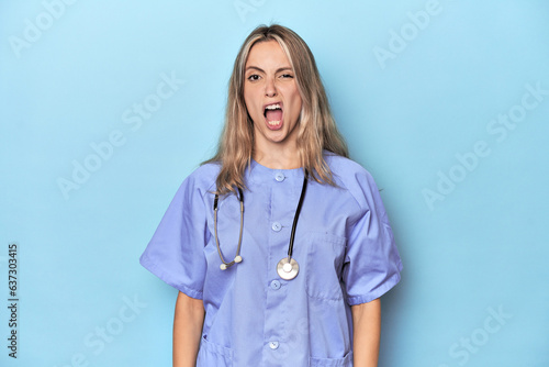 Young caucasian nurse in blue studio screaming very angry and aggressive.