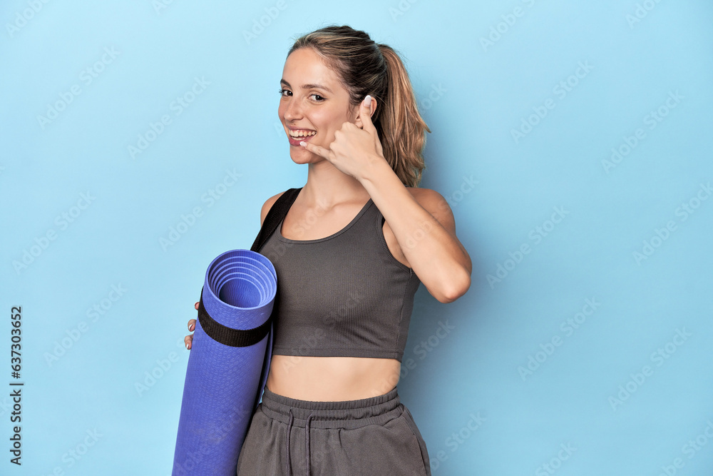 Athlete with a mat in blue studio showing a mobile phone call gesture with fingers. Athlete with a mat in blue studio showing a mobile phone call gesture with fingers.
