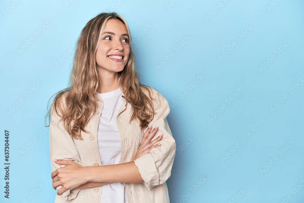 Obraz premium Blonde young caucasian woman in blue studio smiling confident with crossed arms.