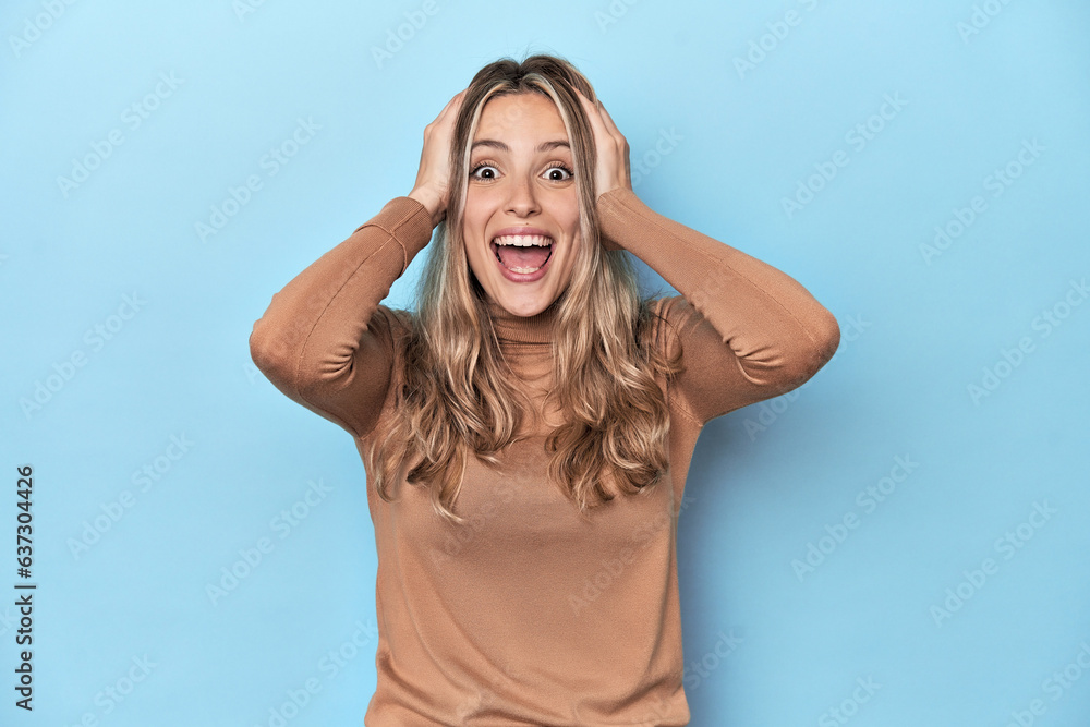 Blonde young caucasian woman in blue studio screaming, very excited, passionate, satisfied with something.