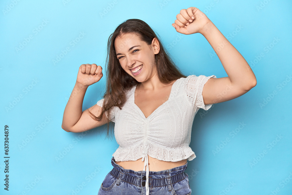 Stylish young woman in white blouse on a blue studio backdrop celebrating a special day, jumps and raise arms with energy. Stylish young woman in white blouse on a blue studio backdrop celebrating a special day, jumps and raise arms with energy.