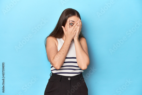Woman in a striped top posing on a blue studio backdrop blink through fingers frightened and nervous.