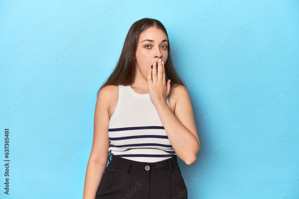 Woman in a striped top posing on a blue studio backdrop yawning showing a tired gesture covering mouth with hand. Woman in a striped top posing on a blue studio backdrop yawning showing a tired gesture covering mouth with hand.