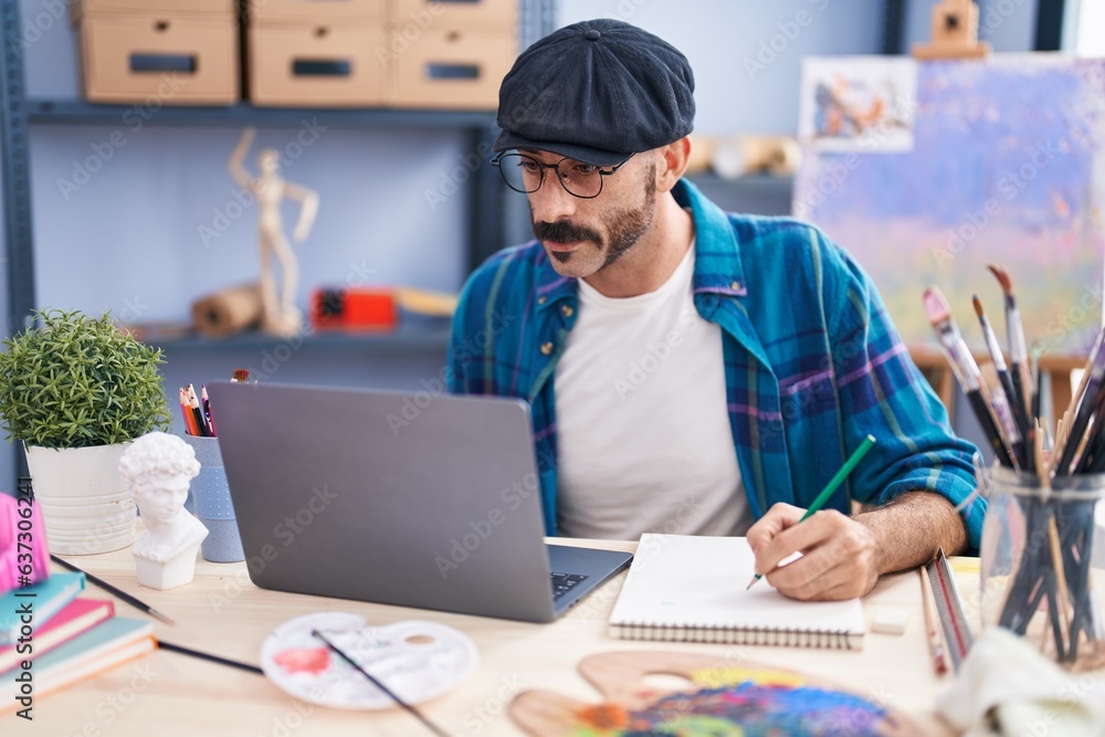 Young hispanic man artist using laptop drawing on notebook at art studio Young hispanic man artist using laptop drawing on notebook at art studio
