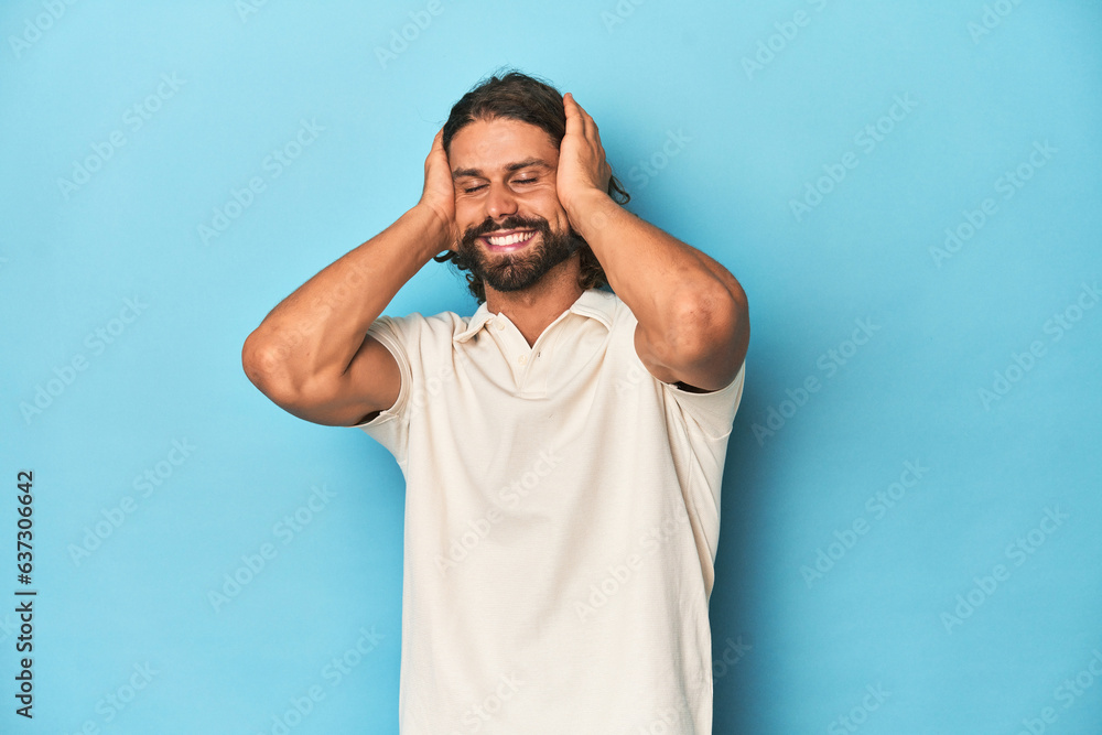 Long-haired man in a white polo, blue studio laughs joyfully keeping hands on head. Happiness concept. Long-haired man in a white polo, blue studio laughs joyfully keeping hands on head. Happiness concept.