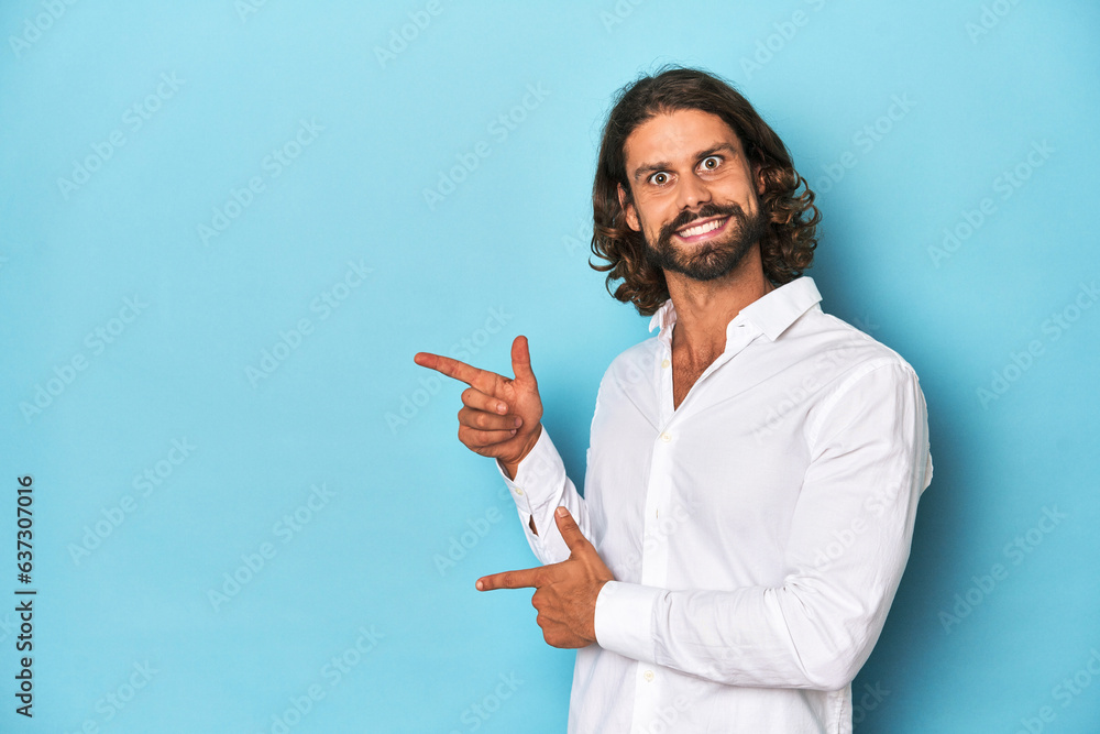 Bearded man in a white shirt, blue backdrop excited pointing with forefingers away. Bearded man in a white shirt, blue backdrop excited pointing with forefingers away.