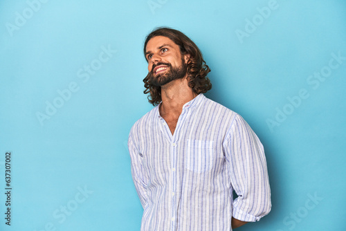 Man with beard in blue striped shirt, blue studio relaxed and happy laughing, neck stretched showing teeth.