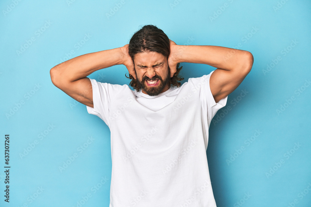 Bearded man in a white shirt, blue backdrop covering ears with hands trying not to hear too loud sound. Bearded man in a white shirt, blue backdrop covering ears with hands trying not to hear too loud sound.