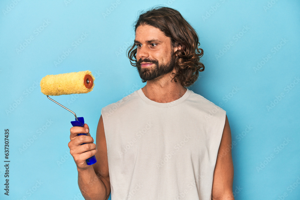 Bearded man holding a paint roller in a blue studio setting. Bearded man holding a paint roller in a blue studio setting.