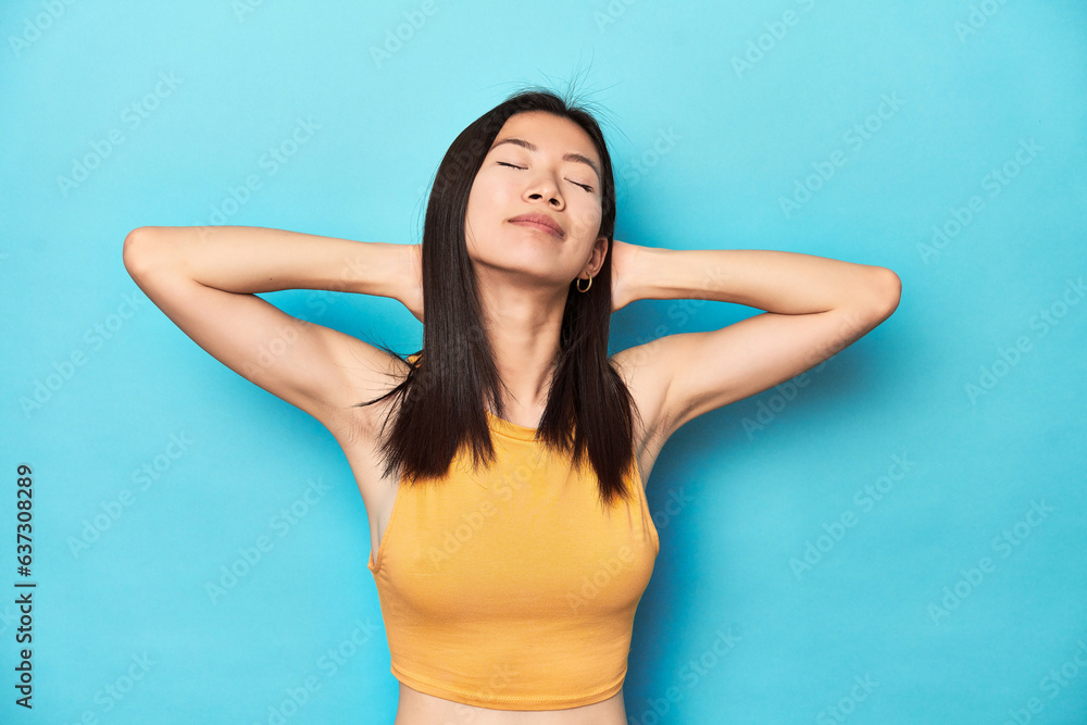 Asian woman in summer yellow top, studio setup, feeling confident, with hands behind the head. Asian woman in summer yellow top, studio setup, feeling confident, with hands behind the head.