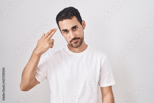 Handsome hispanic man standing over white background shooting and killing oneself pointing hand and fingers to head like gun, suicide gesture.