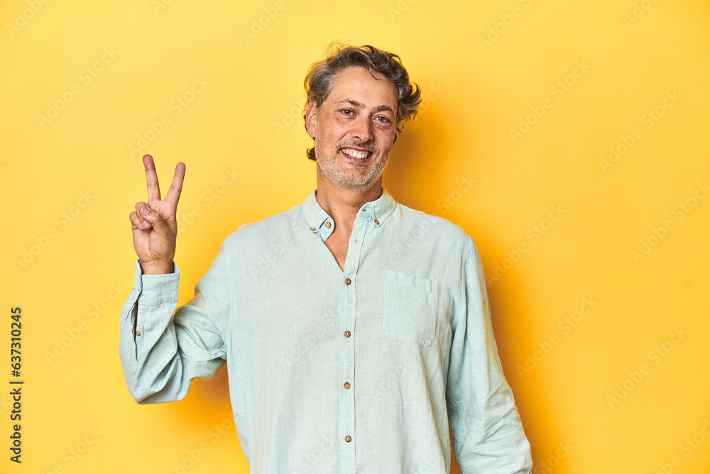 Middle-aged man posing on a yellow backdrop joyful and carefree showing a peace symbol with fingers. Middle-aged man posing on a yellow backdrop joyful and carefree showing a peace symbol with fingers.