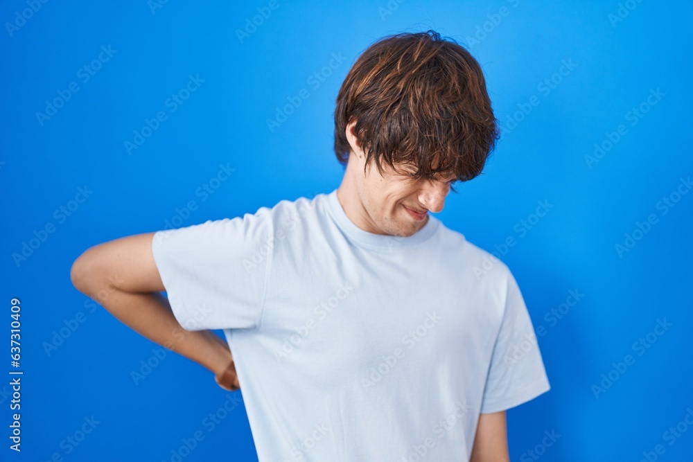 Hispanic young man standing over blue background suffering of backache, touching back with hand, muscular pain Hispanic young man standing over blue background suffering of backache, touching back with hand, muscular pain