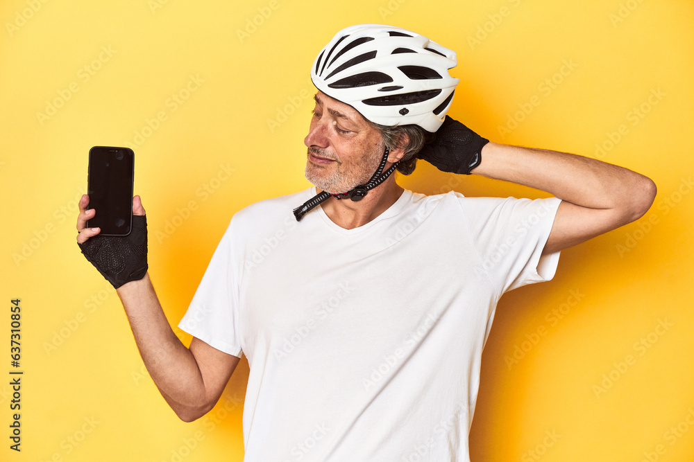 Cyclist man showing phone on yellow backdrop touching back of head, thinking and making a choice. Cyclist man showing phone on yellow backdrop touching back of head, thinking and making a choice.