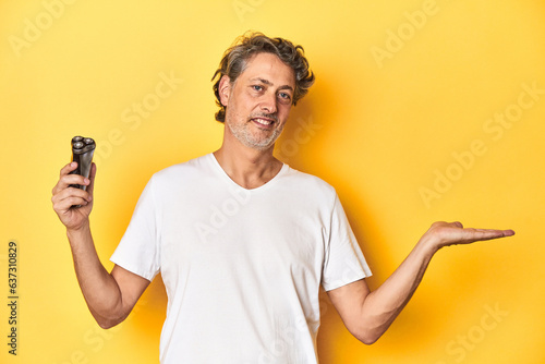 Man holding a razor, yellow studio background showing a copy space on a palm and holding another hand on waist. photo