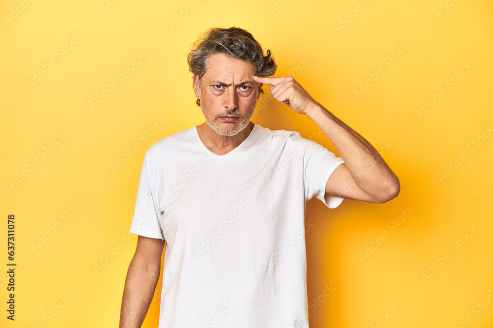 Middle-aged man posing on a yellow backdrop pointing temple with finger, thinking, focused on a task. Middle-aged man posing on a yellow backdrop pointing temple with finger, thinking, focused on a task.