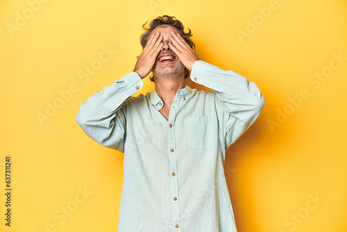 Middle-aged man posing on a yellow backdrop covers eyes with hands, smiles broadly waiting for a surprise.