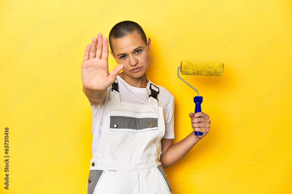 Painter woman, shaved head, with yellow roller in studio standing with outstretched hand showing stop sign, preventing you. Painter woman, shaved head, with yellow roller in studio standing with outstretched hand showing stop sign, preventing you.