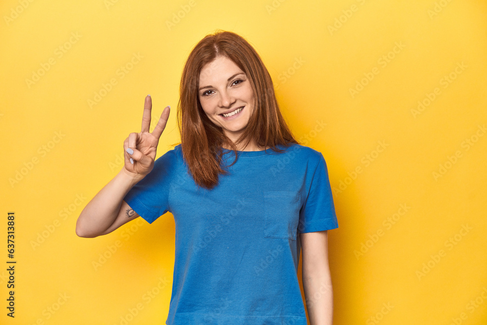 Redhead in blue t-shirt on yellow backdrop joyful and carefree showing a peace symbol with fingers. Redhead in blue t-shirt on yellow backdrop joyful and carefree showing a peace symbol with fingers.