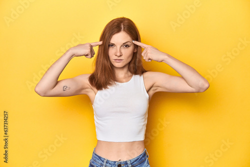 Redhead in white tank top, relaxed studio pose focused on a task, keeping forefingers pointing head.