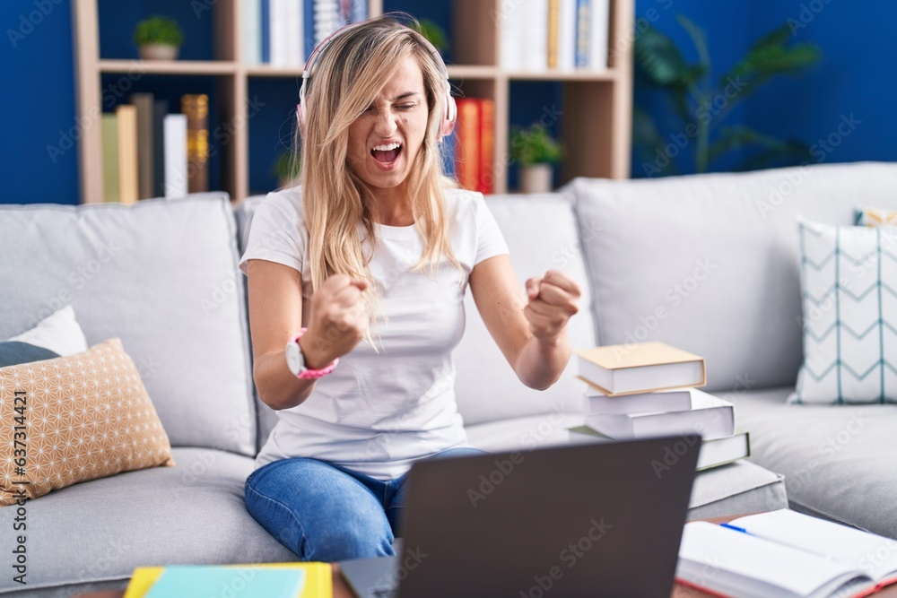Young blonde woman studying using computer laptop at home angry and mad raising fists frustrated and furious while shouting with anger. rage and aggressive concept. Young blonde woman studying using computer laptop at home angry and mad raising fists frustrated and furious while shouting with anger. rage and aggressive concept.