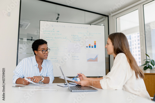 Young enthusiastic African-American female executive in glasses explains new strategy to employee and gesturing. Indoor portrait of diverse work team brainstorming in modern meeting room