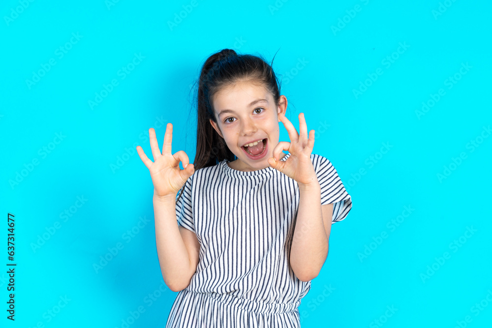 Caucasian kid girl wearing striped dress over blue background showing both hands with fingers in OK sign. Approval or recommending concept Caucasian kid girl wearing striped dress over blue background showing both hands with fingers in OK sign. Approval or recommending concept