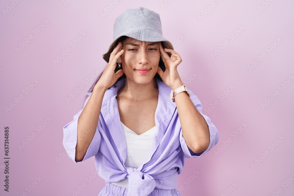 Young hispanic woman standing over pink background wearing hat with hand on head, headache because stress. suffering migraine. Young hispanic woman standing over pink background wearing hat with hand on head, headache because stress. suffering migraine.