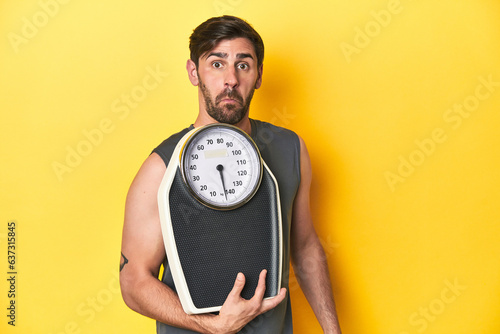 Athletic man with scale  on a yellow studio backdrop shrugs shoulders and open eyes confused.