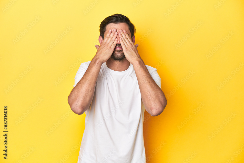 Caucasian man in white t-shirt on yellow studio background afraid covering eyes with hands. Caucasian man in white t-shirt on yellow studio background afraid covering eyes with hands.