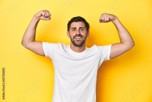 Caucasian man in white t-shirt on yellow studio background showing strength gesture with arms, symbol of feminine power