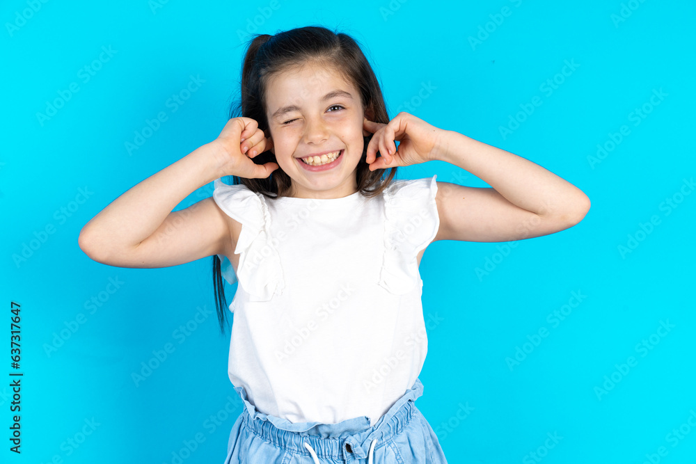 Happy Caucasian kid girl wearing white t-shirt over blue background ignores loud music and plugs ears with fingers asks to turn off sound Happy Caucasian kid girl wearing white t-shirt over blue background ignores loud music and plugs ears with fingers asks to turn off sound
