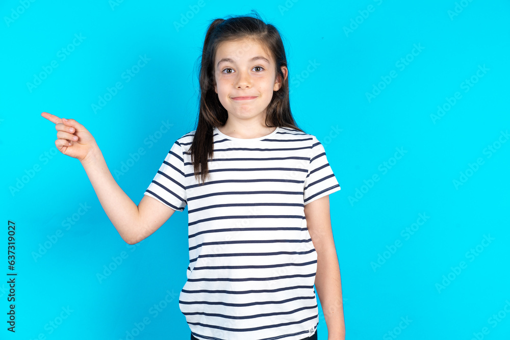 Serious Caucasian kid girl wearing floral dress over blue background smirks face points away on copy space shows something unpleasant. Look at this advertisement. Big price concept Serious Caucasian kid girl wearing floral dress over blue background smirks face points away on copy space shows something unpleasant. Look at this advertisement. Big price concept