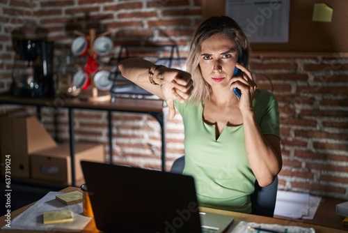 Young beautiful woman working at the office at night speaking on the phone looking unhappy and angry showing rejection and negative with thumbs down gesture. bad expression.