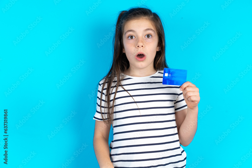 Photo of Caucasian kid girl wearing floral dress over blue background amazed shocked hold credit card payment Photo of Caucasian kid girl wearing floral dress over blue background amazed shocked hold credit card payment
