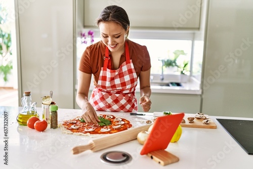 Young beautiful hispanic woman preparing italian pizza watching online recipe on touchpad at the kitchen