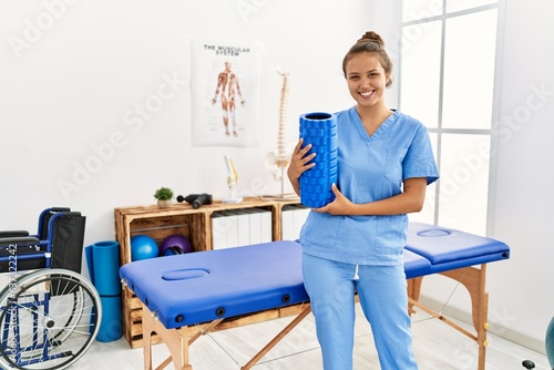 Young beautiful hispanic woman physiotherapist smiling confident holding foam roller at rehab clinic