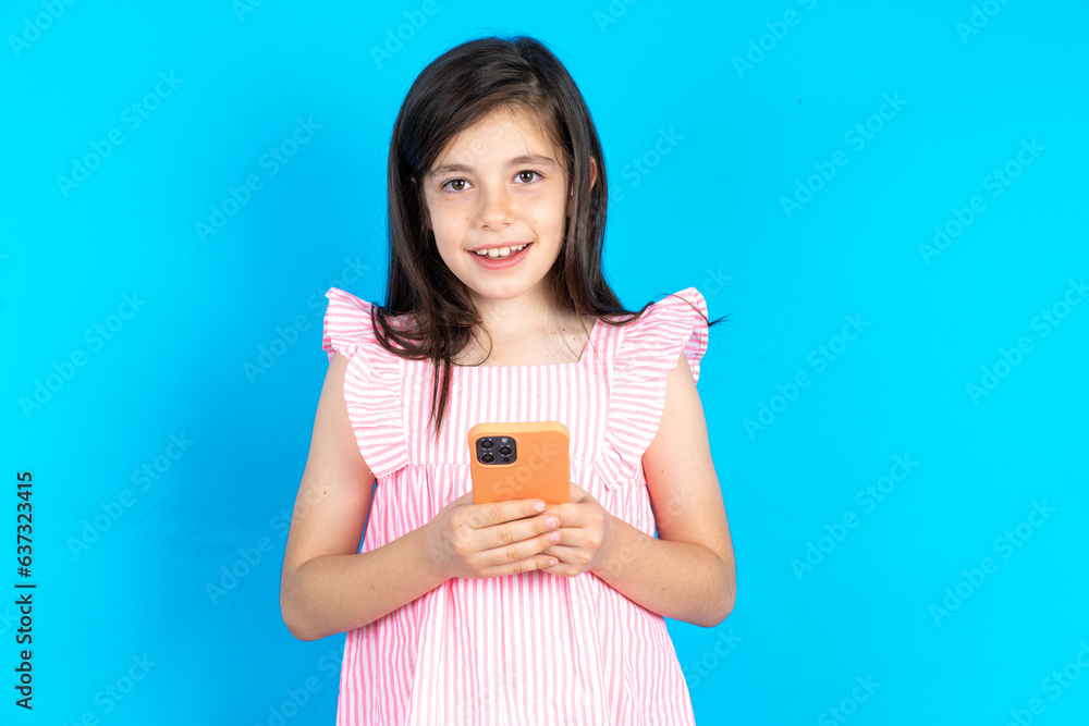 caucaisna kid girl wearing pink dress over blue background enjoys distant communication, uses mobile phone, surfs fast unlimited internet, has pleasant smile, makes shopping online, caucaisna kid girl wearing pink dress over blue background enjoys distant communication, uses mobile phone, surfs fast unlimited internet, has pleasant smile, makes shopping online,