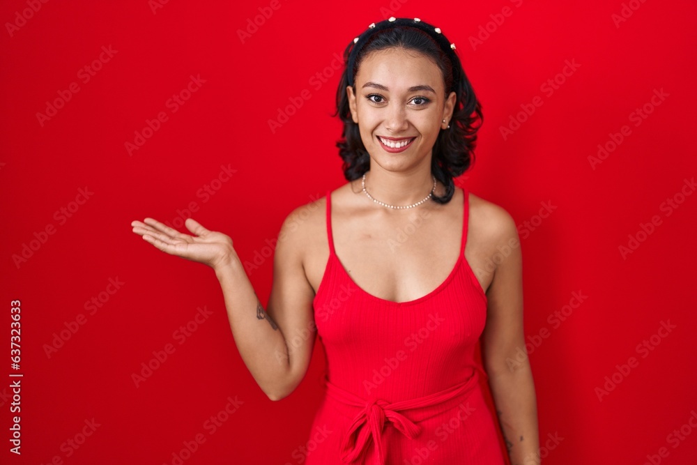 Young hispanic woman standing over red background smiling cheerful presenting and pointing with palm of hand looking at the camera. Young hispanic woman standing over red background smiling cheerful presenting and pointing with palm of hand looking at the camera.
