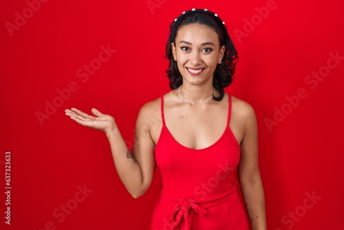 Young hispanic woman standing over red background smiling cheerful presenting and pointing with palm of hand looking at the camera.