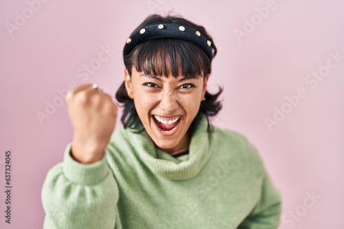 Young beautiful woman standing over pink background angry and mad raising fist frustrated and furious while shouting with anger. rage and aggressive concept.