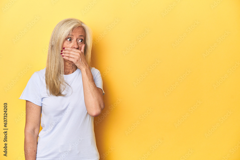 Middle-aged Caucasian woman in white t-shirt, yellow studio thoughtful looking to a copy space covering mouth with hand.