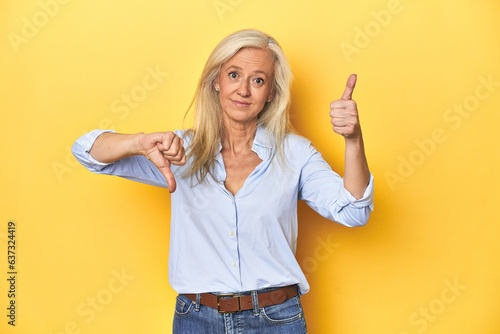 Middle-aged Caucasian woman in blue shirt, yellow studio showing thumbs up and thumbs down, difficult choose concept